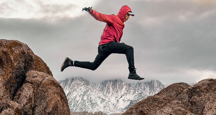 Action shot of a man in a red windbreaker jumping across a ravine 